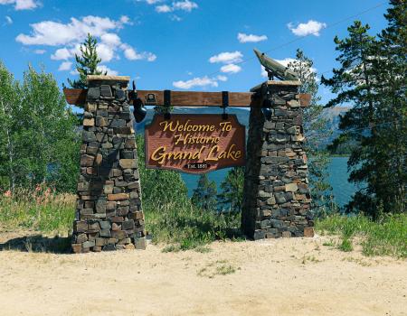 Hanging wooden sign that says "Welcome to Historic Grand Lake" surrounded by trees.